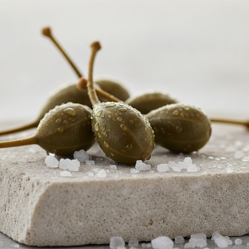 Caper — Fresh green caper berries with scattered salt crystals on pale limestone, morning dew glistening.